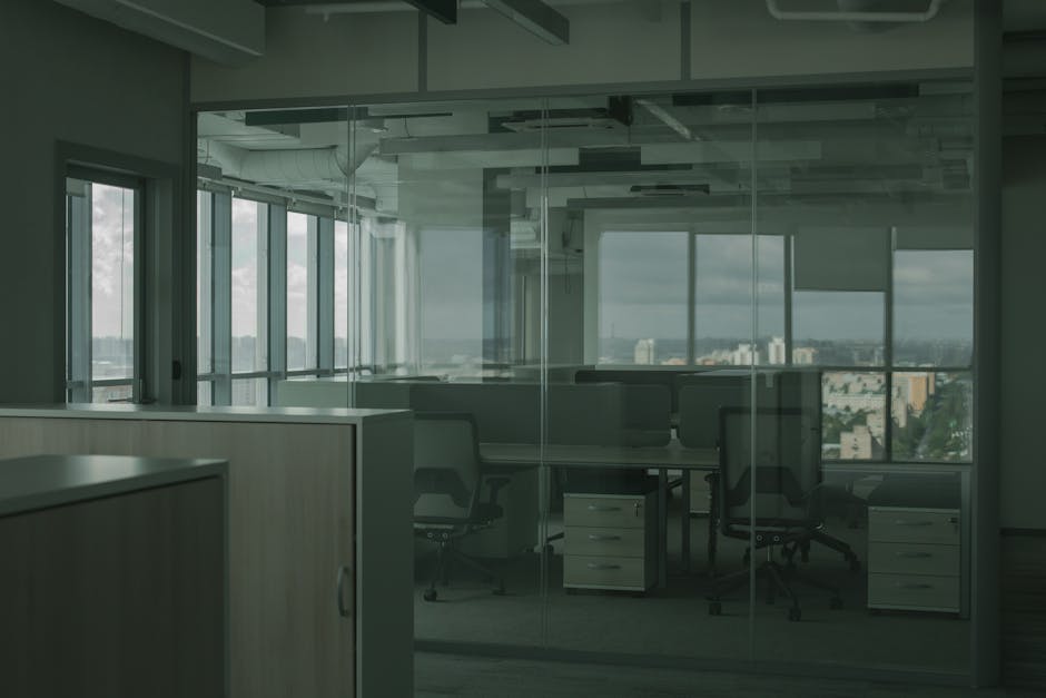A man with dark, curly hair and a beard, wearing an orange long-sleeve shirt, is holding a cardboard box labeled 'OFFICE' in black text. He stands inside a room with a plain, light-colored wall, and a