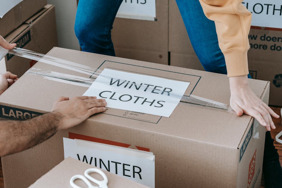 Inside a residential property, a person is seen sealing a large cardboard box labeled 'WINTER CLOTHS' with clear packing tape, indicating the packing process during a home relocation or move. The box 
