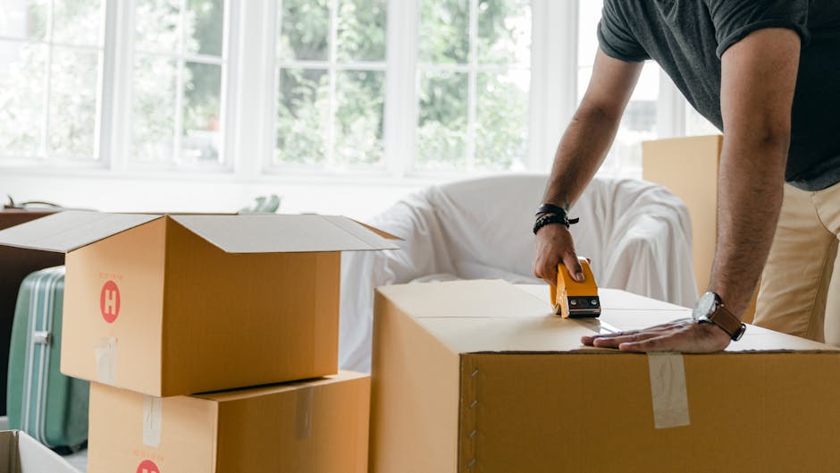 Inside a residential property, a person is seen sealing a large cardboard box labeled 'WINTER CLOTHS' with clear packing tape, indicating the packing process during a home relocation or move. The box 