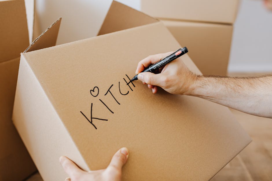 Inside a room near a window with yellow curtains, several cardboard boxes are stacked on a dark-colored piece of furniture. The boxes are sealed with clear packing tape, with one box displaying a red 
